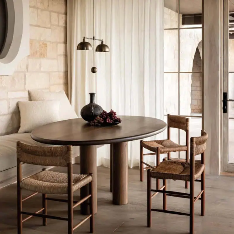A cozy room featuring a round wooden table, woven chairs, and a pendant light. Stone walls and sheer curtains complement the warm, rustic decor.