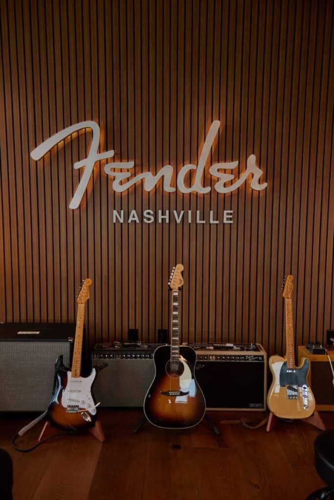 Three guitars displayed in front of a wooden wall with "Fender Nashville" sign, accompanied by amplifiers, creating a musical ambiance.