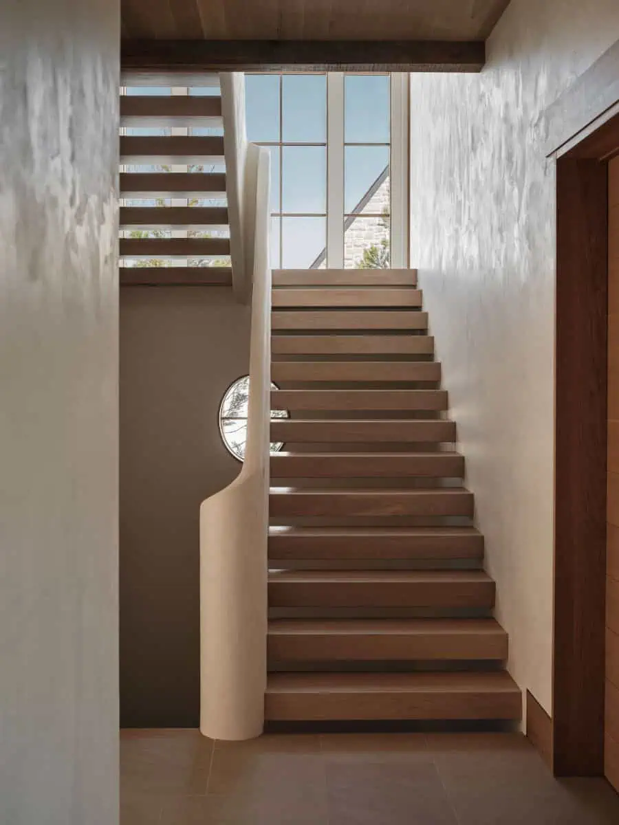 Modern stairway with wooden steps and a round window, illuminated by natural light through large glass panels, creating a minimalist, serene ambiance.