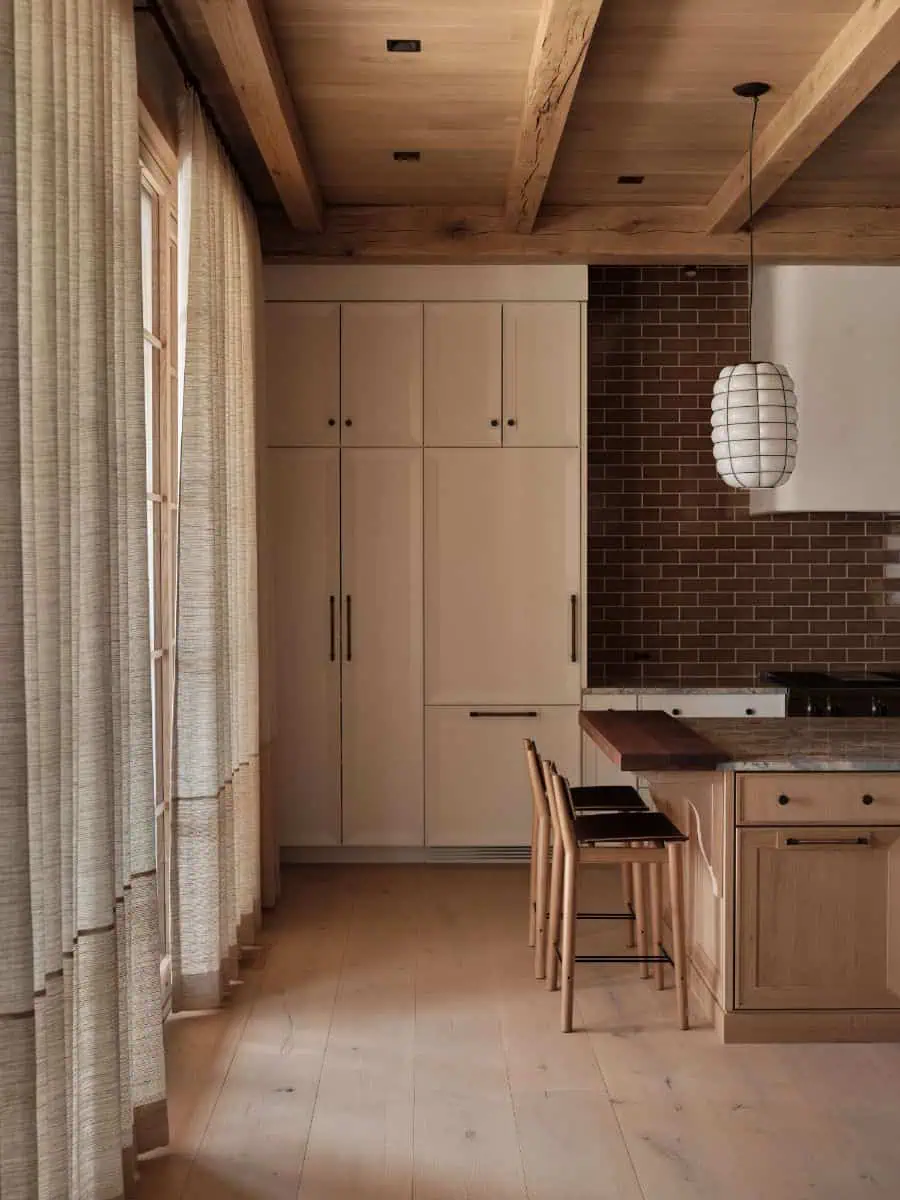 Cozy kitchen with wood beams, brick backsplash, pendant light, and light-colored cabinets. Bar counter with chairs, and textured curtains by windows.