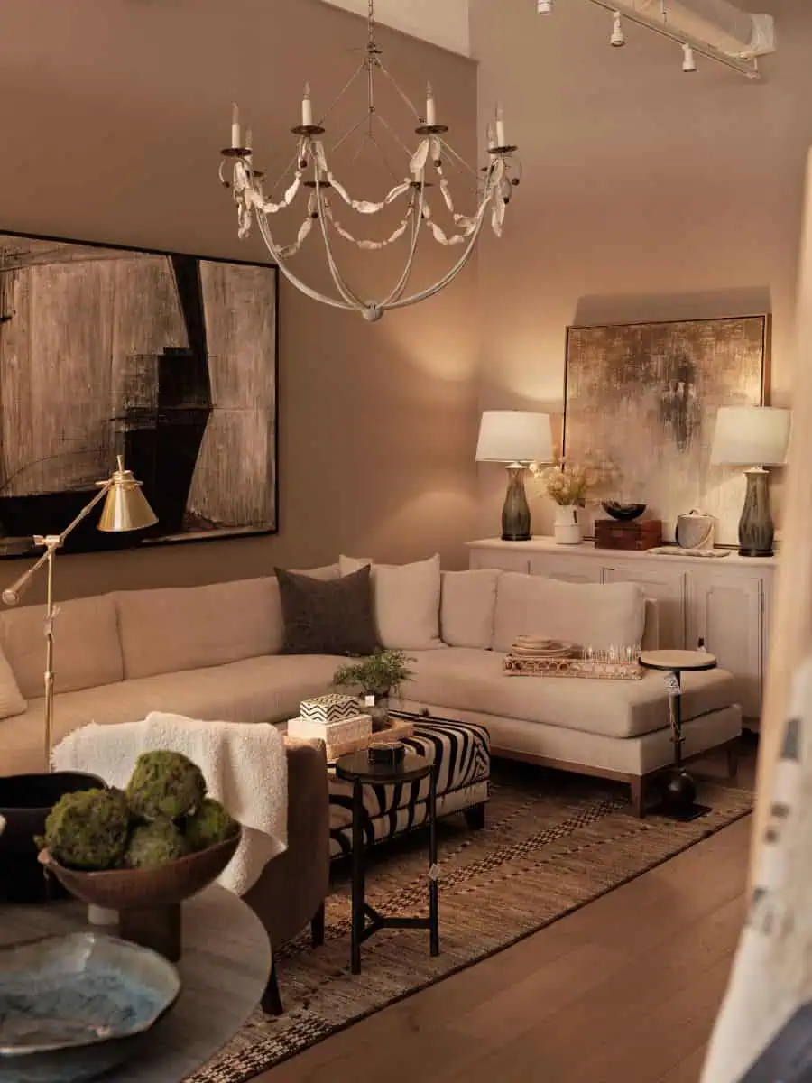 Cozy living room with beige sectional sofa, abstract art, modern chandelier, and ambient lighting. Coffee table accessorized with plants and decorative items.