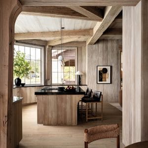 A cozy kitchen features rustic wooden elements, large windows, and a central island with black countertop, surrounded by inviting chairs.