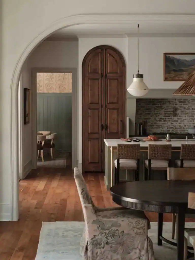 Warm, inviting kitchen and dining area with wood accents, arched doorway, floral-patterned chair, pendant light, and rustic brick backsplash. Peaceful atmosphere.