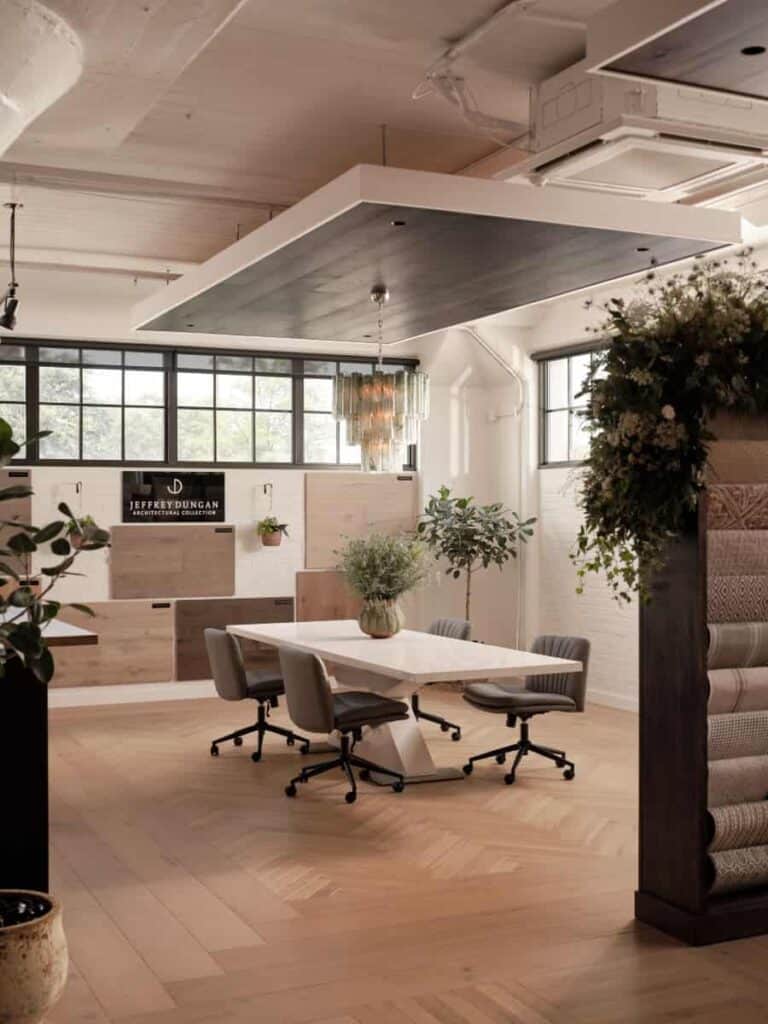 Modern office interior with wooden floors, large windows, and ceiling panels. Central table with chairs, plants, carpets, and lighting fixtures enhance the space.