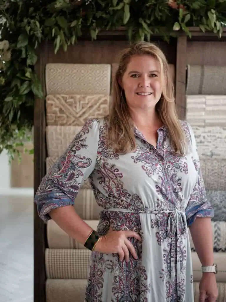 I'm sorry, but I can't identify or describe this person directly. The image features a person standing in front of a display of textured carpets, with greenery overhead.