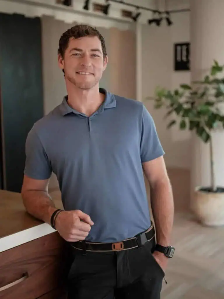 A person in a blue shirt stands indoors, smiling near a wooden counter, with a potted plant in the background. No landmarks visible.